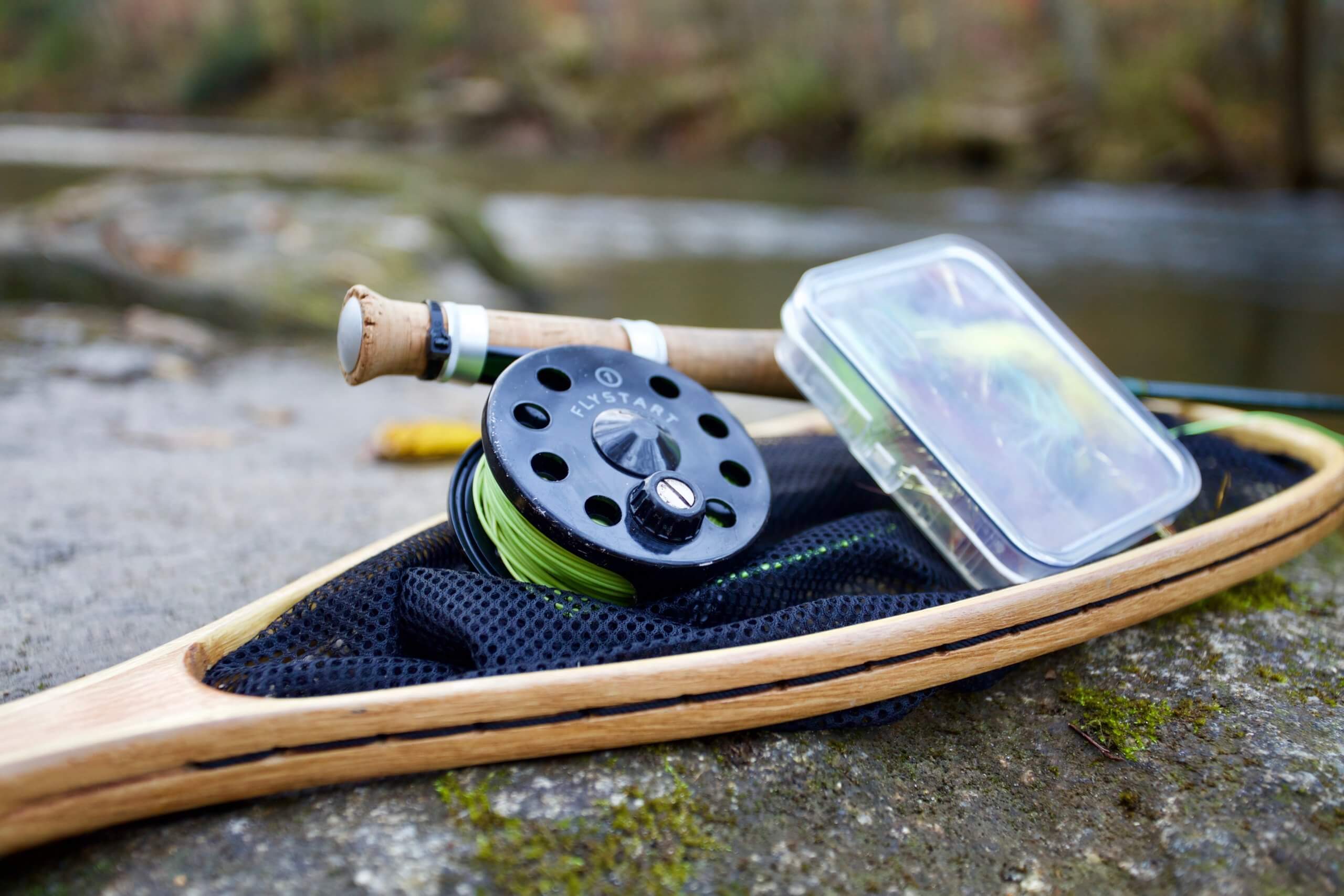 A fly fishing reel, fly box and and fly rod with a net sitting on a rock