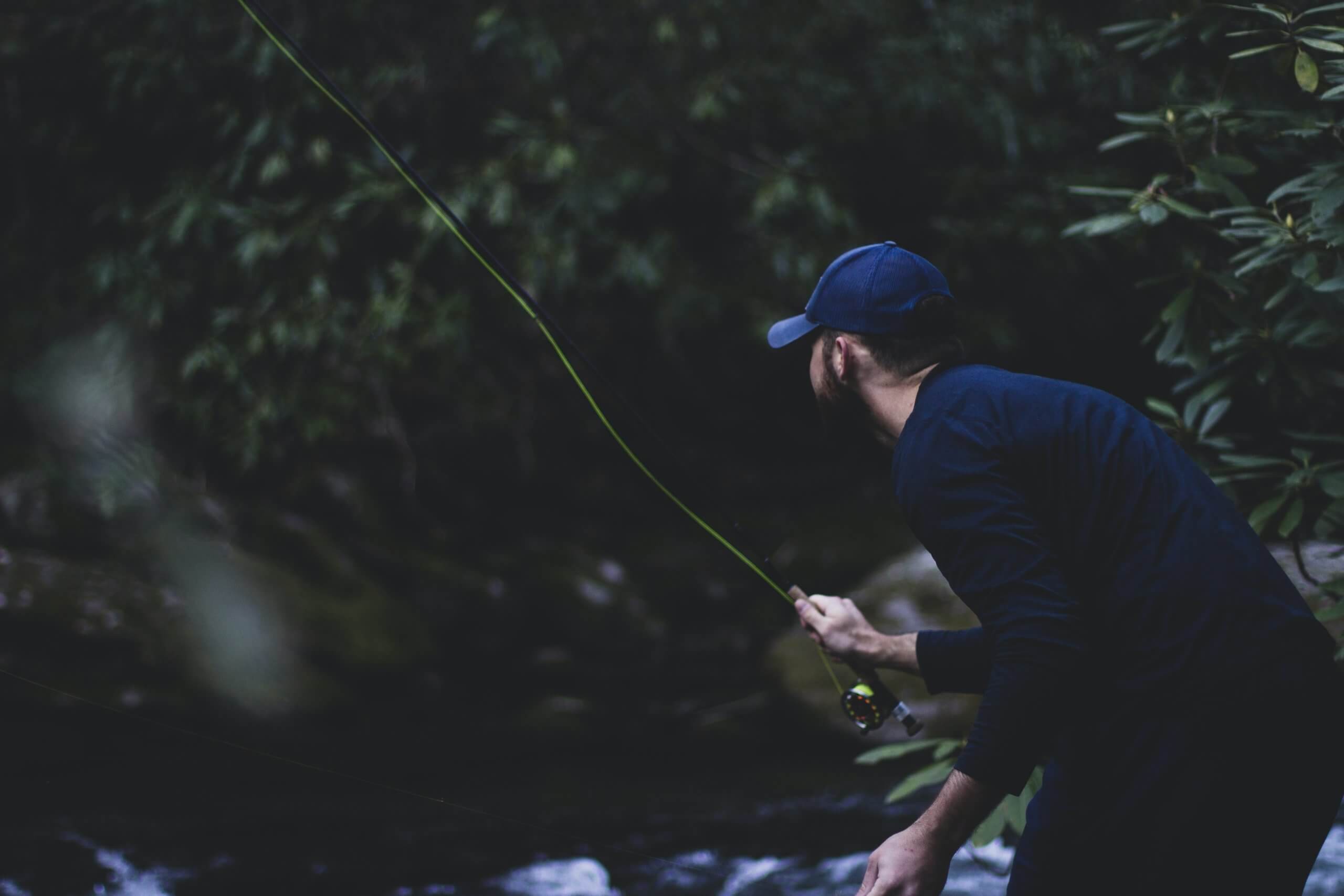 A man in a navy blue shirt and hat casting a fly rod with a dark, green background
